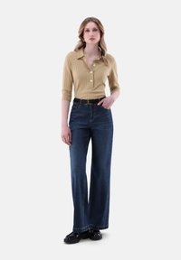 Beige gerippte Poloshirt mit drei Knöpfen, kombiniert mit weiten, dunkelblauen Jeans und schwarzen Lacklederschuhen; schlichtes Design, taillierte Passform.