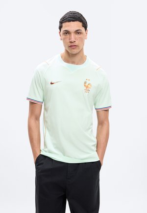 Jeune homme aux cheveux courts et foncés et taches de rousseur, portant un maillot de football France vert menthe clair avec emblème et un pantalon noir, mains dans les poches.