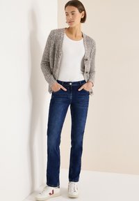 Cárdigan de punto gris texturizado, camiseta blanca ajustada, vaqueros de pierna recta en azul oscuro y zapatillas blancas con detalles en rojo y azul marino.