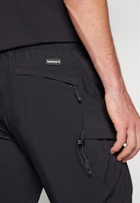 Svarta cargobyxor gjorda av lättviktsmaterial, med en sidoficka med dragkedja och en justerbar dragsko i midjan. Timberland-logotyp synlig.