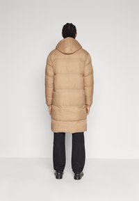 Długa kurtka puchowa w kolorze beżowym z kapturem, z poziomym pikowaniem i lekko zwężanym krojem. Połączona z czarnymi spodniami i obuwiem.