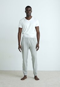 Fehér V nyakú póló és szürke, elasztikus szárú melegítőnadrág. A pólón bal oldalon zseb található; a derékrészen logós részlet látható. Cipő nincs rajta.