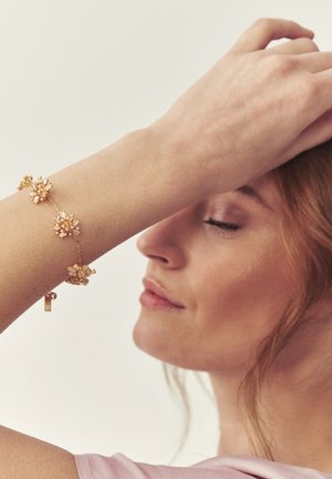 Frau mit geschlossenen Augen, die ein Armband mit goldenen Blumenanhängern trägt und ihren Arm mit der Handfläche an die Stirn legt, vor hellem Hintergrund.