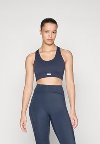 Joven con el cabello recogido, vistiendo un sujetador deportivo azul marino y leggings de tiro alto, de pie contra un fondo claro y sencillo.