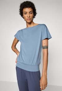 Blue short-sleeve top in lightweight fabric with a relaxed fit, gathered waist, and smooth texture. Paired with navy pants.