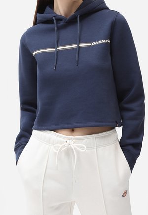 Personne portant un sweat à capuche court bleu marine avec un logo rayé et un pantalon de survêtement blanc à cordon de serrage, doté de poches et d'un petit badge logo.