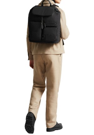Persona vestida con un traje beige y zapatos negros, llevando una mochila negra grande con múltiples bolsillos con cremallera, de espaldas.
