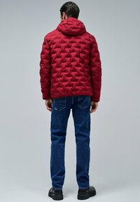 Hombre vistiendo una chaqueta acolchada con capucha roja, jeans azules y botas negras, de pie de espaldas contra un fondo gris liso.