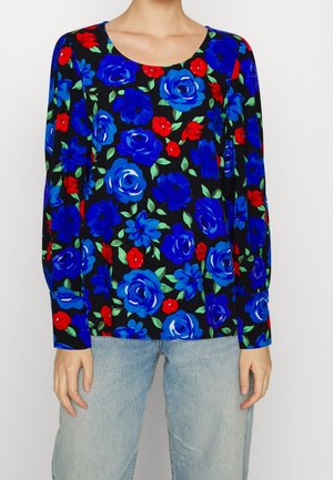 Kvinde iført en sort langærmet bluse med stort blåt og rødt blomsterprint og lyseblå jeans.