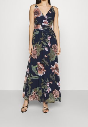 Kvinde iført en ærmeløs, mørkeblå maxikjole med store pink og grønne blomsterprint og sorte høje sandaletter med stropper.