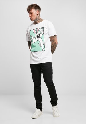 Giovane uomo con tatuaggi al collo e alle braccia, indossa scarpe da ginnastica bianche, pantaloni neri e una t-shirt bianca con stampa del cartone Bugs Bunny.