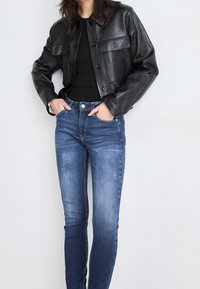 Chaqueta de cuero negra corta con botones en el frente y dos bolsillos en el pecho, combinada con unos jeans ajustados de mezclilla en un lavado azul medio.