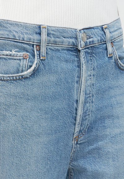 Κοντινό πλάνο ανοιχτόχρωμων τζιν με κουμπωτή μέση, εμπρός τσέπες και θηλιές για ζώνη, φορεμένα με λευκό ριμπ μπλουζάκι.