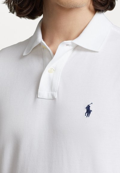 Witte polo met een gestructureerde stof, voorzien van een drieknoopsluiting en een marineblauw geborduurd logo op de linkerborst.
