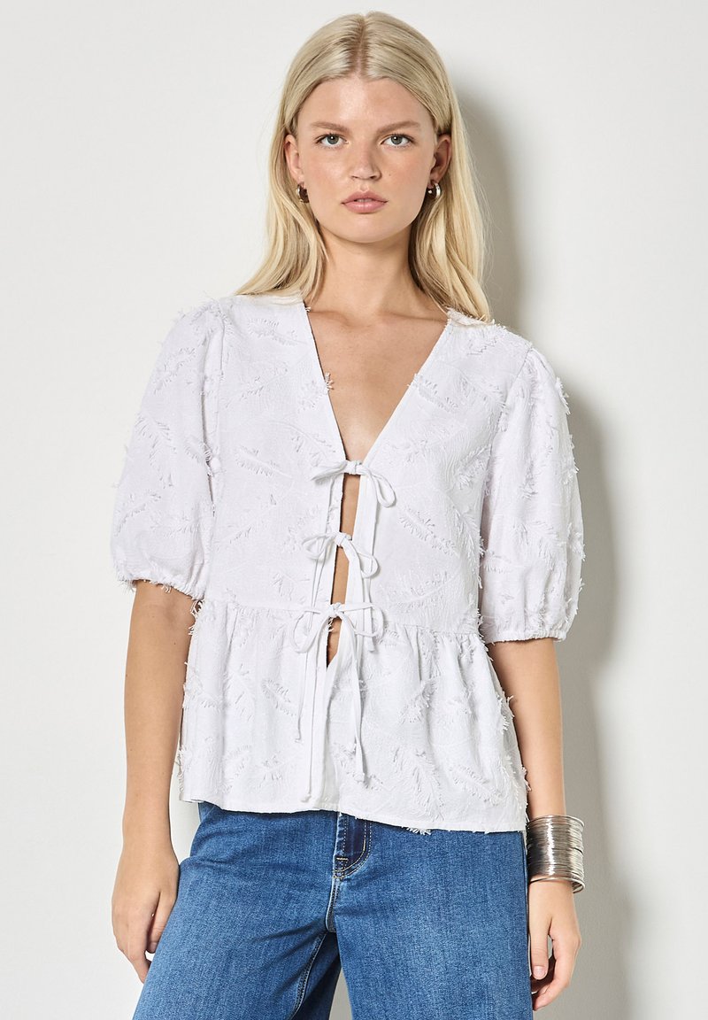 Femme blonde portant un chemisier blanc texturé avec trois liens sur le devant et des manches bouffantes, associé à un jean bleu et un bracelet manchette argenté.