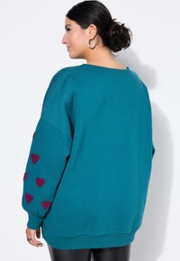 Sudadera oversized en color verde azulado con hombros caídos, que presenta patrones de corazones burdeos en las mangas. Textura suave, cuello redondo y dobladillo acanalado.