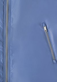 Tissu bleu clair avec deux fermetures éclair en métal argenté, une verticale à gauche et une diagonale à droite, montrant des coutures détaillées.