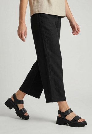 Person, der går iført sorte cropped bukser og sorte chunky hælsandaler med velcro stropper på en ensfarvet baggrund.