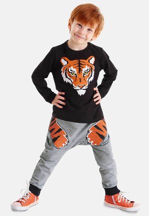 Schwarze Langarmshirt mit einem Tiger-Graphic; graue Hose mit Tigerpfotenabdrücken; orangefarbene Akzente an den Turnschuhen.