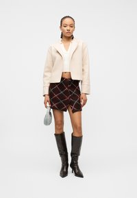 Γυναίκα με πλεκτό μαλλί που φοράει κρεμ μπουφάν και κοντό τοπ, σκούρα καρό μίνι φούστα, μαύρες μπότες μέχρι το γόνατο, κρατώντας μικρή ανοιχτό μπλε τσάντα.