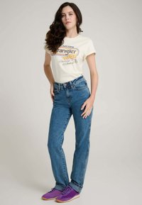 Cremefarbenes T-Shirt mit Retro-Aufdruck, blaue hoch taillierte Jeans und lila Sportschuhe. Das Outfit besticht durch eine lockere Passform und einen lässigen Stil.