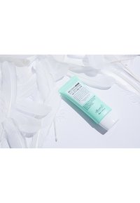 Grön mintfärgad solkräm i tub med vit flipplock, omgiven av vita fjädrar på en ljus bakgrund, märkt med produktinformation.