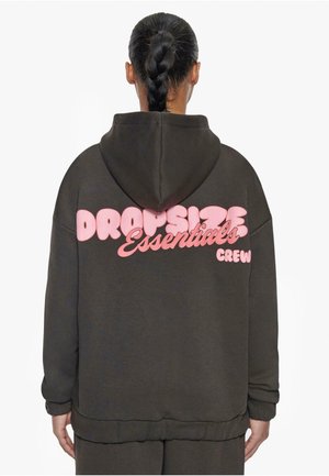 Osoba z warkoczykami, ubrana w ciemną bluzę z kapturem z różowym napisem "DROPSIZE Essentials CREW" na plecach na białym tle.