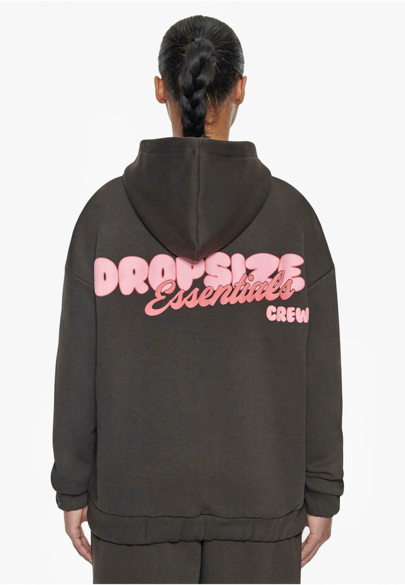 Personne aux cheveux tressés portant un sweat à capuche sombre avec un texte rose « DROPSIZE Essentials CREW » dans le dos sur fond blanc.
