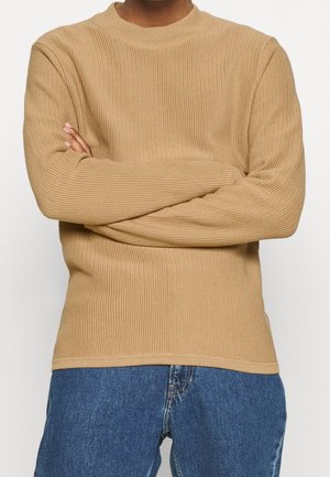 Personne portant un pull en maille côtelée beige, les bras croisés, associé à un jean bleu, sur un fond blanc uni.