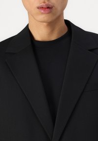 Schwarzer Blazer mit scharfen Revers über einem schwarzen Rundhalsoberteil, das eine glatte Textur und ein minimalistisches Design aufweist, das sich auf klare Linien konzentriert.