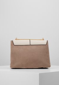 Bolso de ante marrón con una solapa crema, cadena dorada y costuras negras. Textura suave y forma rectangular con un diseño limpio.