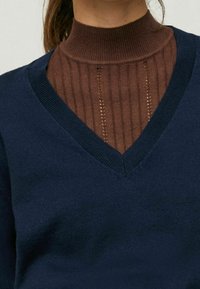 Marineblauer V-Ausschnitt-Pullover mit feiner Stricktextur über einem braunen, gerippten Rollkragenpullover mit vertikalen Linien und dezenten Lochdetails.