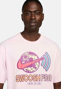 Camiseta de algodón rosa claro con un gráfico colorido que presenta un globo terráqueo, olas y el texto "SWOOSH FM". Mangas cortas y corte relajado.