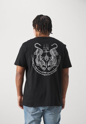 Mężczyzna z warkoczami, ubrany w czarną koszulkę z tyłu z dwoma białymi tygrysami i napisami oraz jasnoniebieskie dżinsy.