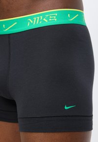 Boxers Nike noirs avec taille verte et logo Nike swoosh vert sur la cuisse gauche, portés par une personne.