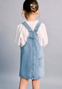 Denimové šaty na ramínka v světle modré barvě s nastavitelnými popruhy, jednou zadní kapsou s motivem "Hello Kitty" a pod nimi texturované bílé tričko s dlouhým rukávem.