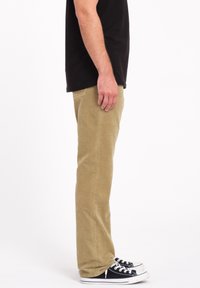 Chinos beige de corte recto, exhibidos en un maniquí de lado, combinados con zapatillas negras y blancas. Textura de tela suave.