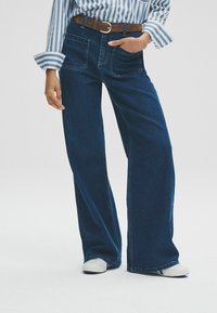 Dunkelblaue Jeans im Schlagstil mit zwei Fronttaschen, detaillierter Nähte und einem braunen Gürtel. Kombiniert mit einem gestreiften Hemd und weißen Schuhen.