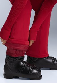Fekete síbakancs három állítható csattal, piros párnázott nadrággal viselve, bordázott mandzsettákkal és elasztikus, sima felülettel.