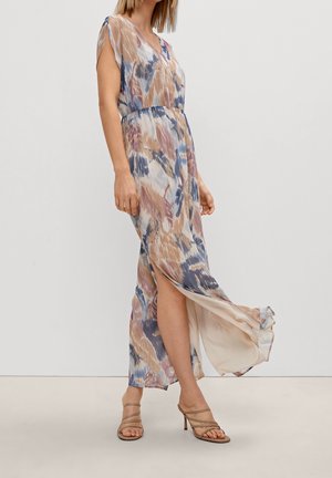 Kvinde iført en flydende, ærmeløs maxikjole med V-hals i pastelabstrakt print og sidespalte, kombineret med beige høje sandaletter med stropper.