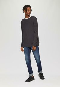 Dunkelgrauer gerippter Pullover mit rundem Halsausschnitt und seitlichen Schlitzen, kombiniert mit blauen Jeans und schwarzen Sportschuhen, alles vor einem neutralen Hintergrund.