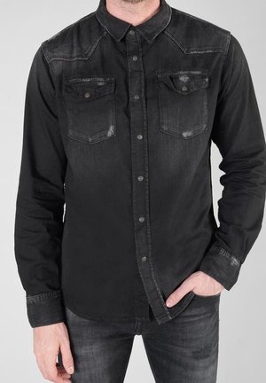 Mann trägt schwarzes Denimhemd mit geknöpften Brusttaschen, eine Hand in der Tasche, vor hellgrauem Hintergrund.