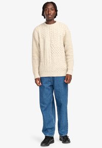 Lichtbeige kabelgebreide trui met een ronde hals en geribbelde manchetten, gecombineerd met blauwe denim jeans en zwarte schoenen. Standaard pasvorm.
