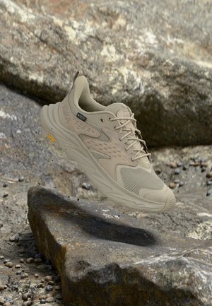 Beiger Wanderschuh mit Mesh- und Wildlederdetails schwebt über felsigem Gelände mit kleinen Steinen und großen, strukturierten Felsen.
