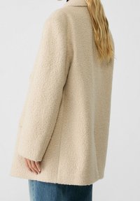 Beige structuurjas met een zachte, pluizige oppervlakte, voorzien van een kraag en lange mouwen, gecombineerd met lichtblauwe jeans.