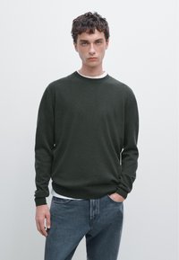 Donkergroene gebreide trui met een ronde hals, lange mouwen en een losse pasvorm, gecombineerd met blauwe denim jeans.