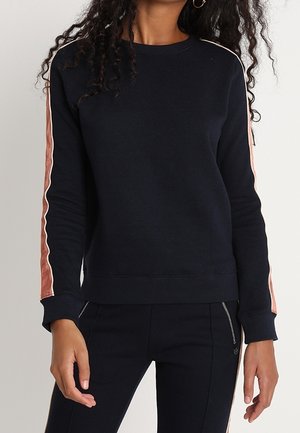 Femme portant un pull noir à manches longues avec des rayures roses et blanches le long des manches, associé à un pantalon noir avec des détails zippés.