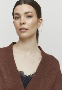 Brązowy sweter z owijanym krojem, wykonany z miękkiego materiału, z głębokim dekoltem w serek, noszony na czarnej koronkowej bluzce. Gładka struktura bez widocznych wzorów.