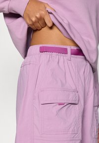 Ανοιχτό μωβ cargo σορτς με υφασμάτινη υφή, ελαστική μέση και ροζ ζώνη ως διακοσμητικό στοιχείο. Διαθέτει μεγάλες πλαϊνές τσέπες.