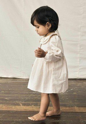 Licht beige gestreepte jurk met een ruches kraag en pofmouwen, gedragen door een kind, dat blootsvoets op houten vloerdelen staat.